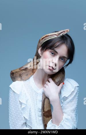 Ritratto di una giovane donna elegante in abito bianco, serpente drappeggiato sulle spalle, sguardo intenso, posa su sfondo blu-grigio. Foto Stock