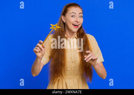 Donna adulta maga strega che gestisce con bastone magico, facendo avverare il desiderio, lanciando incantesimi maghi, pubblicando sconti per le vacanze. Redhead girl isolata su sfondo blu Foto Stock