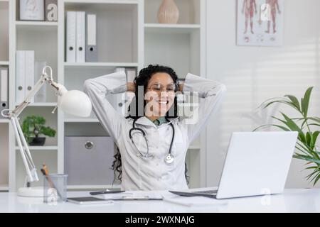 Gioiosa dottoressa latina in cappotto bianco che fa una pausa alla scrivania dell'ufficio con il laptop, sentendosi felice e contenta. Foto Stock