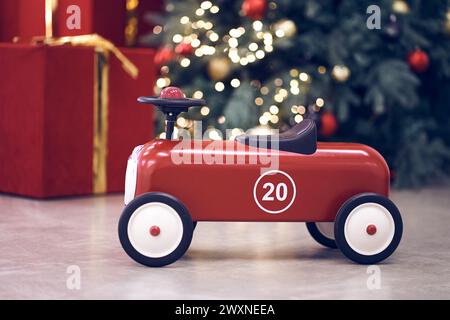 Vista dell'auto retrò rossa per bambini sullo sfondo di regali in scatole rosse con archi dorati, ghirlanda decorata con alberi di Natale, luci a corde, palline rosse e blu. Sfondo, spazio copia, bokeh. Foto Stock