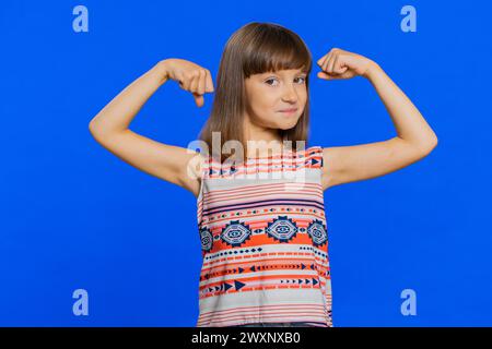 Sono forte e in salute. Giovane ragazza carina che mostra bicipiti e sembra fiduciosa sentendo la forza di lottare per i diritti, l'energia per ottenere il successo vince. Bambino bruna preteen su sfondo blu Foto Stock