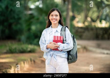 Giovane donna radiosa con abiti bianchi e uno zaino grigio con un libro rosso Foto Stock