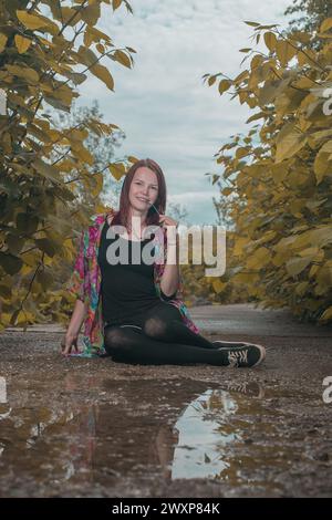 Una giovane donna con i capelli rossi morde gli occhiali da sole e posa mentre si siede su un sentiero pieno di foglie intorno a lei. Donna circondata da verdi lussureggianti Foto Stock