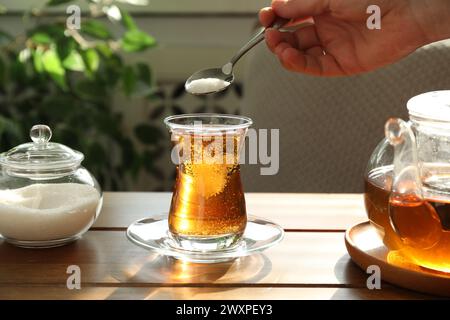 Donna che aggiunge zucchero nel tè aromatico al tavolo di legno all'interno, primo piano Foto Stock