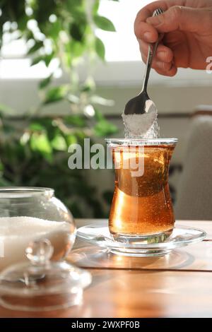 Donna che aggiunge zucchero nel tè aromatico al tavolo di legno all'interno, primo piano Foto Stock