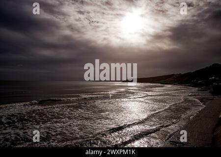 La luce del sole irradia attraverso nuvole grigie piovose e brilla attraverso le onde che corrono su una spiaggia, Scarborough South Bay, North Yorkshire, Inghilterra. Foto Stock