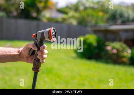 Tubo da giardino con ugello regolabile. Mano dell'uomo che tiene la pistola a spruzzo e innaffia le piante, spruzzando acqua sull'erba nel cortile. Foto Stock