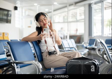 Una bella e rilassata passeggero turistica asiatica si sta godendo la musica con le sue cuffie nel terminal dell'aeroporto mentre aspetta il suo volo a bordo Foto Stock