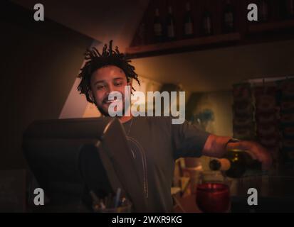 Giovane barista afroamericano che porge vino rosso in bicchiere e sorride Foto Stock