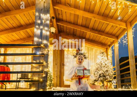 Una donna con un vestito bianco che tiene una scatola regalo davanti a un albero di Natale. Foto Stock