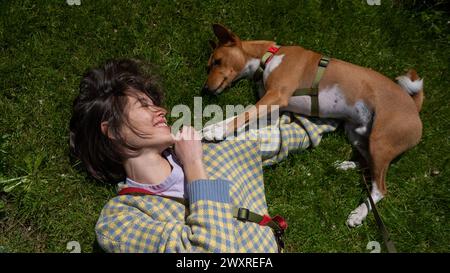 Vista dall'alto di una bella giovane donna e di un cane basenji africano sdraiato sull'erba verde. Foto Stock