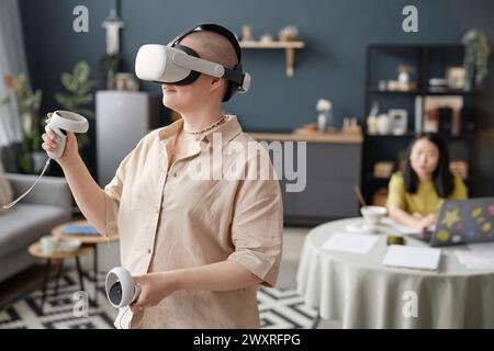 Foto selettiva di una giovane donna caucasica che indossa un visore che gioca a un videogame VR, la sua amica che studia in background Foto Stock