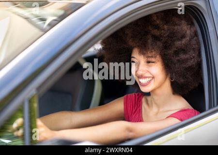 Sorridente eccitata giovane donna etnica con auto di guida dai capelli afro scuri il giorno d'estate in città Foto Stock