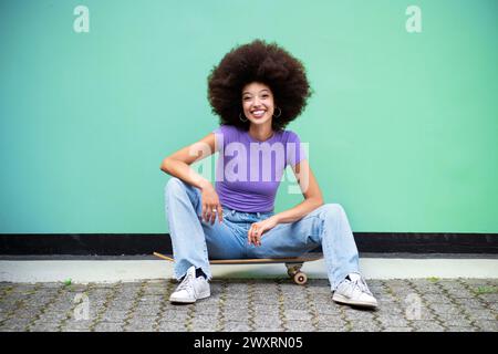 Corpo completo di giovane e allegra donna marocchina con acconciatura afro e abiti casual che guardano la macchina fotografica mentre si siede alla luce del giorno sullo skateboard in via Foto Stock