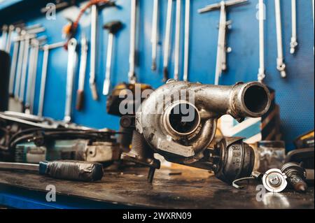 Primo piano del motore turbo per auto insieme ai pezzi di ricambio in assistenza in officina e posizionati sul banco di riparazione contro gli attrezzi e gli inserti di bulloneria appesi Foto Stock