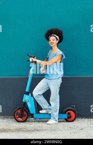 Corpo pieno di giovani donne etniche sorridenti con capelli afro che indossano un abito in denim con scooter ecologico per strada Foto Stock