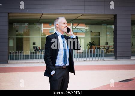 un uomo d'affari sorridente che parla al telefono sul suo smartphone all'aperto. Foto Stock