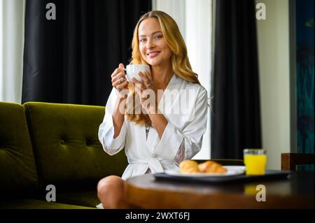 Una donna sorridente in accappatoio si rilassa con il suo caffè mattutino su un morbido divano verde Foto Stock