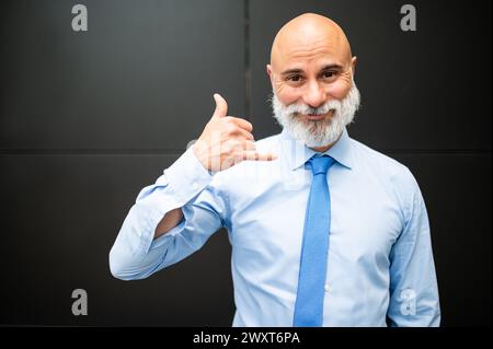 Ritratto uomo d'affari maturo, calvo ed elegante, con barba bianca all'aperto che fa il segnale di chiamata con la mano Foto Stock