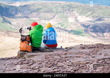 Brecon Beacons, Galles. Un camminatore maschio e una femmina siedono sulla cresta del Corn Du alla sua sommità. L'uomo sta usando una macchina fotografica per scattare una foto dall'alto. Foto Stock