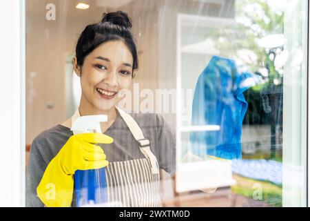 Giovane donna in grembiule e guanti gialli che puliscono finestre Foto Stock