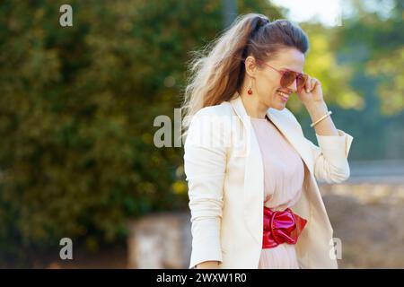 donna alla moda felice in abito rosa e giacca bianca in città con occhiali da sole. Foto Stock