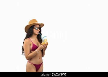 La bellissima donna latina si rilassa sulla spiaggia sorseggiando un cocktail tropicale e indossando un elegante cappello da sole, che incarna l'essenza di un perfetto Foto Stock