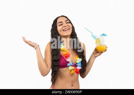 La giovane e gioiosa latina che indossa un bikini ride allegramente mentre tiene un cocktail estivo, adornata da una collana di fiori intorno al collo, su un sole Foto Stock