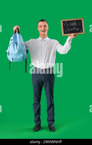 Allegro studente maschio che tiene la lavagna con testo SCUOLA ADDIO su sfondo verde. Concetto di fine scuola Foto Stock