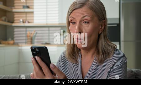 Sorpreso felice anziano 60s donna caucasica leggere buone notizie utilizzando il telefono cellulare sul divano a casa il preavviso bancario ha stupito la pensionessa donna che vince online Foto Stock