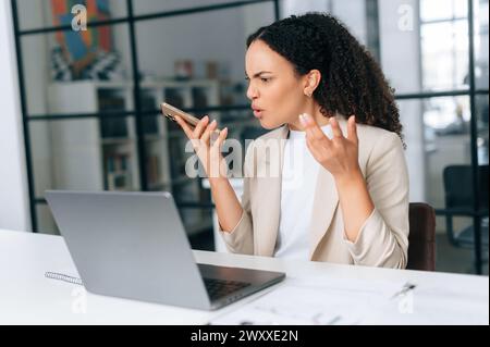 Arrabbiata, infastidita, brasiliana o ispanica, curly businesswoman, responsabile finanziario aziendale, siede in un ufficio moderno, parla irritabilmente su un telefono cellulare con l'altoparlante o registra un messaggio audio Foto Stock