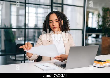 Donna brasiliana riccissima o latina concentrata in abiti eleganti, dipendente, segretaria o responsabile finanziario, siede sul posto di lavoro con un notebook nell'ufficio moderno, studiando documenti e analizzando Foto Stock