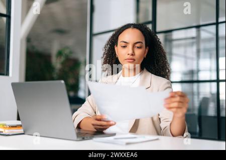 Donna brasiliana o latina concentrata in abiti eleganti, dipendente, segretaria o responsabile finanziario, siede sul posto di lavoro con un notebook nell'ufficio moderno, studia documenti e analizza Foto Stock