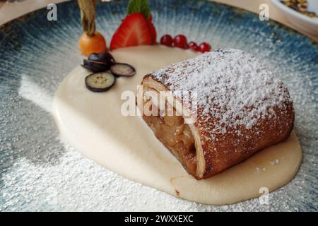 Delizioso strudel di mele spolverato con zucchero a velo, servito su un sofisticato piatto di ceramica blu con un turbinio di salsa alla vaniglia. Foto Stock