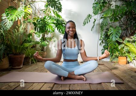 Donna nera soddisfatta in posizione di loto, sorridente in senso lato, rilassandosi nello studio di yoga nella giungla urbana. Foto Stock