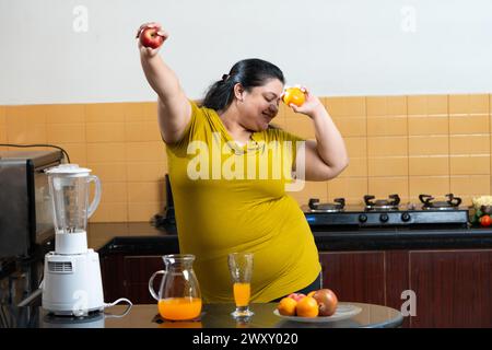 Donna indiana grassa in sovrappeso che si eccita mentre tiene in mano mela e arancia in una cucina. Concetto di mangiare sano. Foto Stock