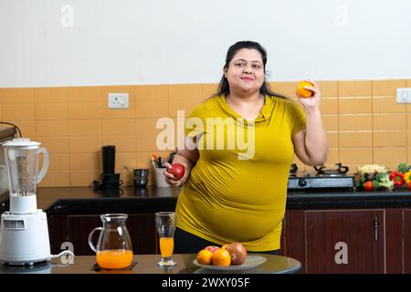 Donna indiana grassa in sovrappeso sorridente, guardando la macchina fotografica mentre tiene in mano mela e arancia in una cucina. Concetto di mangiare sano. Foto Stock