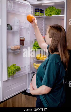 Giovane donna incinta con un'arancia vicino al frigorifero di casa. Mangiare sano. Foto Stock