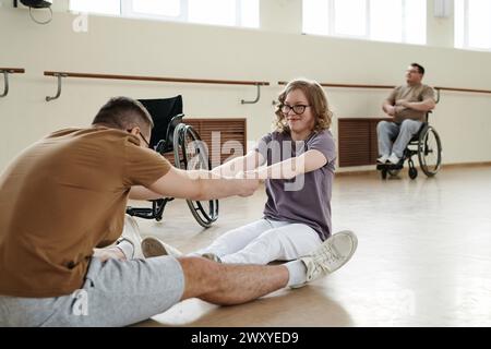 Giovane donna gioiosa con disabilità seduta sul pavimento a fare esercizio di stretching insieme al suo compagno di ballo Foto Stock