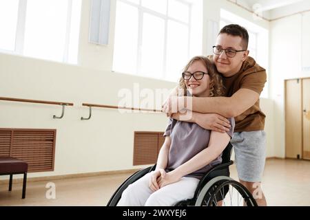Colpo medio lungo di un giovane amorevole che abbraccia una giovane donna felice con disabilità in uno studio di danza in sedia a rotelle Foto Stock