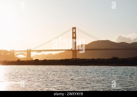 Il sole al tramonto proietta una silhouette dorata del Golden Gate Bridge, creando una scena serena sulla baia con sfondi di montagna nebbiosi Foto Stock