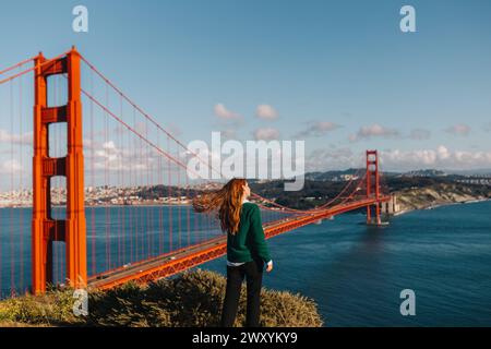 La donna con vista sul retro è stupita mentre il vento cattura i suoi capelli, affacciato sul magnifico Golden Gate Bridge in una vibrante giornata di primavera Foto Stock