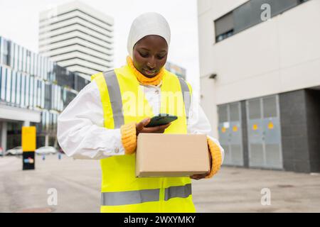 Una donna musulmana africana sorridente che indossa un hijab e un giubbotto di sicurezza contiene un pacchetto e utilizza il suo smartphone in un ambiente urbano. Foto Stock