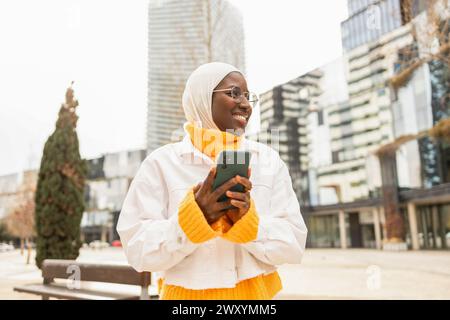 Una allegra donna africana in un hijab bianco e una vivace sciarpa gialla ha uno smartphone, godendosi il suo tempo in un paesaggio urbano moderno. Foto Stock