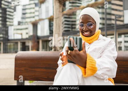 Allegra donna afroamericana che indossa un hijab e occhiali, guarda lo schermo dello smartphone con un sorriso luminoso, seduta su una panchina all'aperto con un arredo urbano Foto Stock