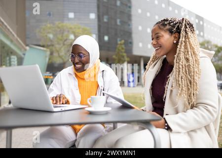 Due donne afroamericane, una che indossa un hijab, stanno conducendo una piacevole conversazione in un bar all'aperto con un portatile sul tavolo, simboleggiando Mu Foto Stock