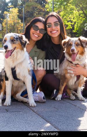 Due giovani donne felici che si inginocchiano e posano con i loro allegri cani da pastore australiani in un parco soleggiato Foto Stock