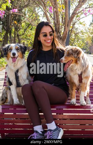 Una donna allegra che indossa gli occhiali da sole siede su una panchina del parco fiancheggiata da un paio di cani da pastore australiani felici Foto Stock