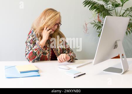 Una donna d'affari bionda e concentrata parla al telefono mentre utilizza un computer alla scrivania dell'ufficio, con uno stabilimento in background Foto Stock
