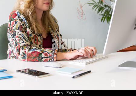 Una donna d'affari bionda professionista si concentra sulla digitazione su una tastiera, lavorando diligentemente nel suo ufficio ordinato e organizzato Foto Stock